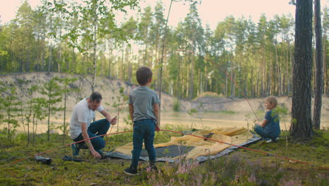 The-young-man-and-two-young-boys-together-set-up-a-tent-camp-for-the-night-during-the-campaign.-Fatherhood-and-a-happy-childhood.-Father-and-sons-put-up-a-tent-together-in-the-woods